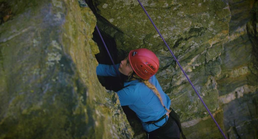 A young person wearing safety gear is secured by ropes as they navigate a crack in a rock wall they are climbing. 